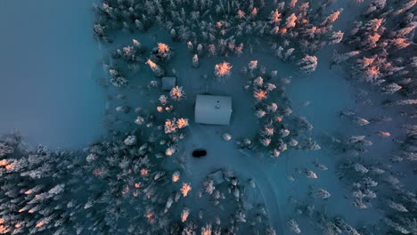 Top-down-drone-shot-above-a-cabin-in-middle-of-snowy-forest,-sunrise-in-Lapland