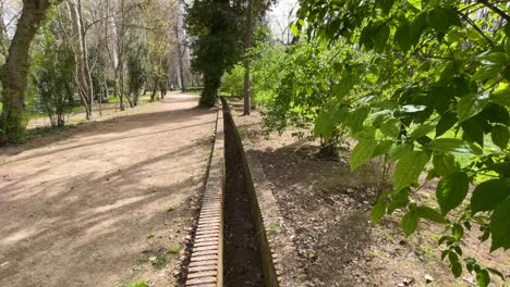 Wir-Sehen-Die-Aufnahmen-Eines-Gemauerten-Bewässerungskanals-Auf-Einem-Sandweg,-Auf-Der-Einen-Seite-Treiben-Bäume-Ihre-Ersten-Knospen-Aus-Und-Auf-Der-Anderen-Seite-Grüne-Blätter-Im-Jardin-Del-Principe-Aranjuez,-Spanien