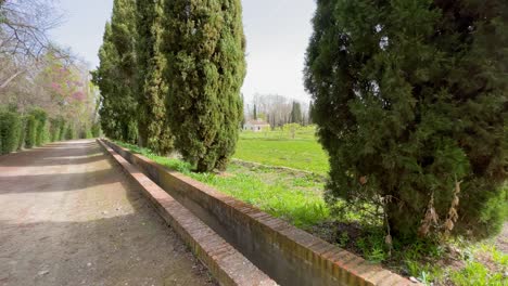 We-see-in-the-Jardin-del-Principe-in-Aranjuez-an-extension-of-trees-with-a-farmhouse-and-a-dirt-road-with-cypresses-on-one-side-and-on-the-other-side-hedges-and-trees-there-is-an-irrigation-canal