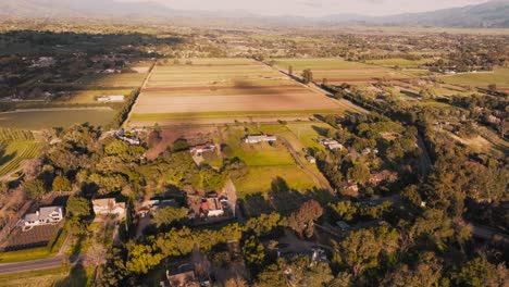 Aerial-Footage-of-Santa-Ynez-Homes-and-Farms-at-Golden-Hour,-Green-Fields-and-Crops-with-Mountains-on-Horizon
