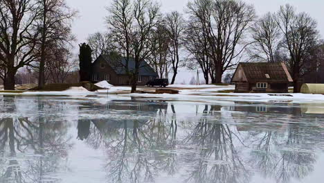 Winter-scenery-of-thaw-puddle,-rustic-houses-in-reflection,-amazing-view-of-Latvia-countryside