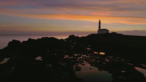Aerial-sunset-panoramic-landscape-in-Menorca-coastline-Lighthouse-skyline-golden-colorful-sky-with-pink-tones,-Island,-sea-shore-rocky-environment