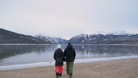 Pareja-Tomados-De-La-Mano-Caminando-Hasta-El-Borde-De-Un-Lago-Congelado-Con-Montañas-Cubiertas-De-Nieve-En-El-Fondo,-Estático