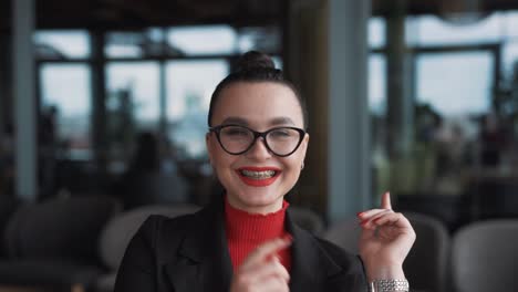 portrait-features-a-beautiful-young-woman-in-glasses-and-braces,-a-freelancer,-sitting-in-a-stylish-restaurant,-dressed-in-business-attire,-smiling,-and-pointing-at-her-braces