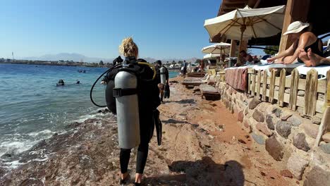 Blonde-woman-walks-in-Egyptian-beach-after-scuba-diving-with-umbrellas-bars-along-sea-coastline-in-daylight-as-seen-from-behind