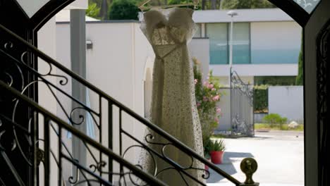 A-white-wedding-dress-hanging-on-its-hanger-at-the-entrance-to-a-magnificent-villa,-we-see-in-the-foreground-the-wrought-iron-banisters-of-the-staircase