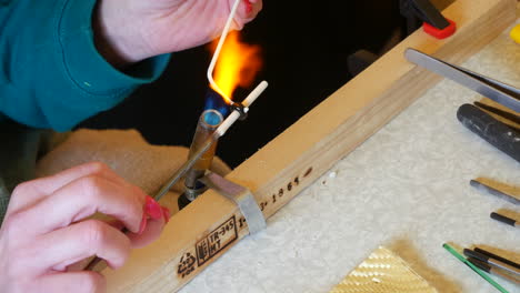 Close-up-of-hands-using-a-blowtorch-to-heat-glass-rods-adding-decoration-in-a-jewellery-making-process,-with-tools-on-a-workbench