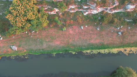 Herbstliche-Pracht,-Luftaufnahme-Von-Lebendigen-Bäumen,-In-Leuchtenden-Farbtönen-Von-Rot,-Orange,-Gold-Und-Grün,-Säumen-Das-Ruhige-Flussufer-Darunter-Und-Kleine-Weiße-Felsige-Moosbedeckte-Klippen