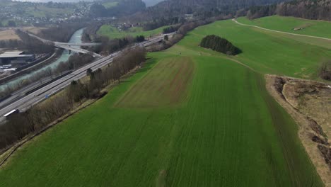 Drone-footage-of-Highway-next-to-green-Field-and-River-with-Forest-and-Snow-covered-Mountain-Landscape-in-Background