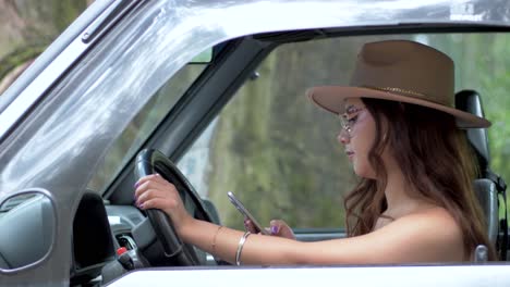 Die-Moderne-Eleganz-Einer-Lächelnden-Ecuadorianerin,-Die-Auf-Dem-Fahrersitz-Sitzt-Und-Mit-Brille-Und-Modischem-Hut-Ihr-Mobiltelefon-Benutzt