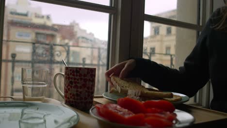 Frau-Im-Pyjama-Bereitet-Frühstück-Am-Fenster-Vor-Und-Arrangiert-Brot-Und-Tomaten-Auf-Dem-Tisch