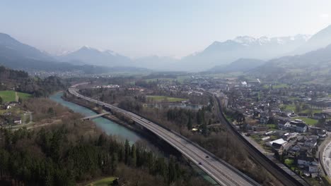 Aerial-view-of-Cars-driving-on-Highway-next-to-a-river-with-River-with-snow-covered-mountains-in-background