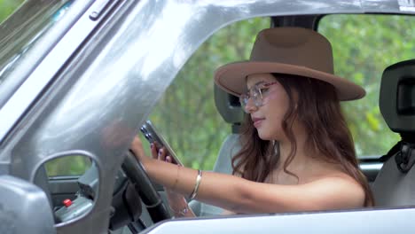 A-woman-in-glasses-and-hat-smiling-while-using-her-mobile-phone-in-the-driver's-seat-of-a-car