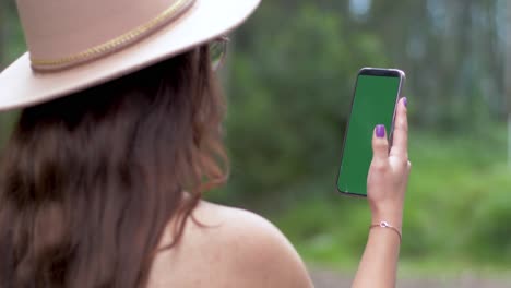 A-girl-with-an-open-hair,-wearing-a-hat-and-glasses,-engages-with-her-smartphone-against-a-backdrop-of-lush-green-trees-and-grass