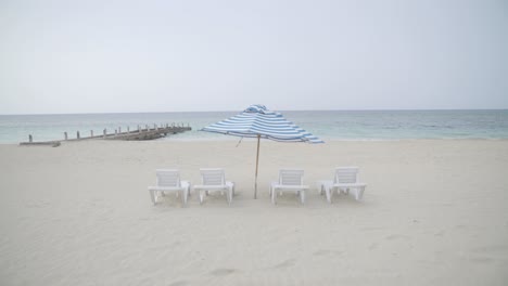 Blau-weißer-Sonnenschirm-Am-Windigen-Einsamen-Strand-Mit-Weißem-Tropischen-Sand-Und-Grauem-Himmel,-Etabliert