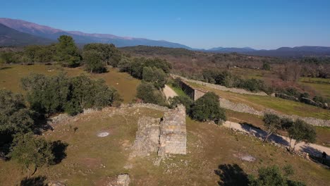 Drone-flight-in-reverse-with-a-medieval-ruined-hermitage-from-the-11th-century-surrounded-by-fields-with-pastures-and-groves,-in-the-background-the-mountains-of-the-Sierra-de-Gredos-in-Avila-Spain
