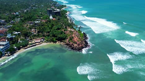 Aerial-drone-of-Coconut-tree-hill-bay-palm-plantation-on-headland-coastline-with-local-town-village-Indian-ocean-waves-Mirissa-Point-Sri-Lanka-Asia