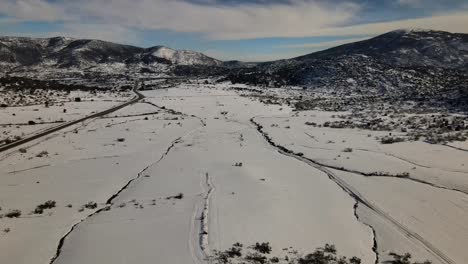 drone-flight-on-a-snowy-plain-seeing-grooves-in-the-snow-created-by-water-currents-and-there-is-a-road-on-one-side-without-vehicles-with-a-background-of-mountains-in-winter-in-Avila-Spain