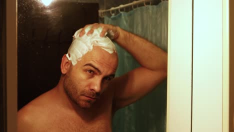 moody-shot-of-a-bald-bearded-man,-applying-shaving-foam-to-his-head,-preparing-it-for-a-shave,-in-front-of-the-bathroom-mirror