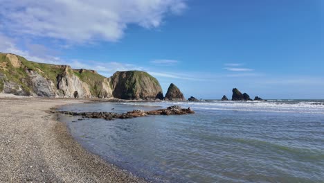 Crystal-clear-water-gentle-waves-on-pebble-beach-with-sea-cliffs-and-blue-sky-with-puffy-wihite-clouds-bright-spring-day-Copper-Coast-Waterford-Ireland