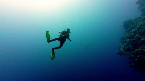 Scuba-Diver-Flota-En-Medio-De-Un-Acantilado-Submarino-Azul-Respirando-Burbujas-En-Dahab,-Egipto,-Luz-Solar-Panorámica-Sobre-El-Océano