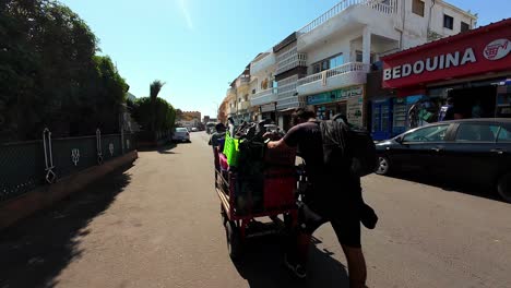 Scuba-divers-in-Dahab,-Egypt-with-Tank-Trolley-Dive-Gear-walking-down-a-street-lined-with-shops-and-parked-cars-in-sunny,-urban-area-with-buildings-featuring-balconies-and-Arabic-signage-Backpackers