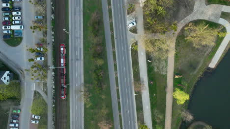 Aerial-view-of-a-red-tram-moving-along-a-track-parallel-to-a-tree-lined-road,-with-a-canal-visible-to-the-right