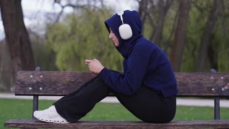 Frau-Hört-Musik-über-Kopfhörer-Und-Spielt-Spiele-Auf-Dem-Smartphone,-Sitzt-Auf-Einer-Parkbank