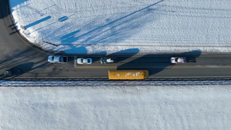 Toma-Aérea-De-Seguimiento-De-Arriba-Hacia-Abajo-De-Un-Autobús-Escolar-Amarillo-Conduciendo-Por-Un-Camino-Rural-Rodeado-De-Un-Paisaje-Cubierto-De-Nieve-En-Invierno