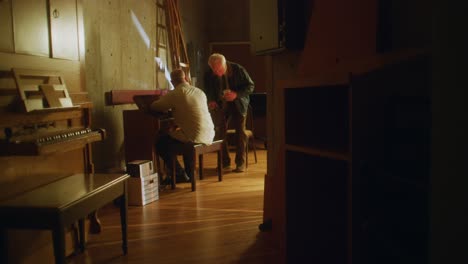 Man-sits-at-piano-in-hallway-and-is-joined-by-a-friend
