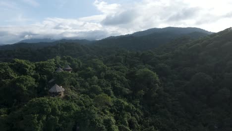 Eine-Drohne-Schwenkt-Von-Den-Prähistorischen-Felsbrocken-Am-Strand-Nach-Oben-Und-Gibt-Den-Blick-Auf-Die-Berge-Und-Den-Regenwald-Von-Tayrona-In-Kolumbien-Frei,-Während-Im-Hintergrund-Die-Sonne-Untergeht