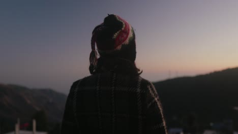 Girl-with-cap-and-winter-wears-standing-and-looking-away-at-the-horizon-in-evening---from-the-back-POV-shot