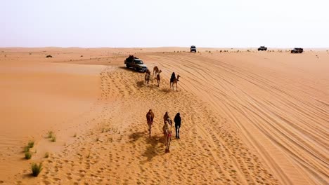 Drone-shot-over-a-cavalcade-of-camels,-cars-and-people,-group-of-Arabs-riding-on-the-Arabian-desert,-in-Saudi-Arabia---dolly,-aerial-view
