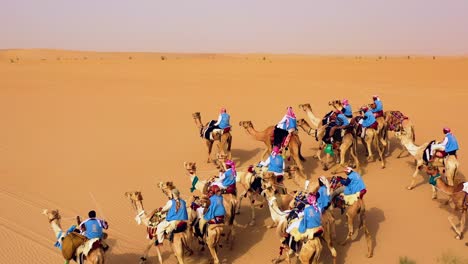 Aerial-view-around-a-camel-cavalcade-riding-over-the-Arabian-desert,-sunny-day,-in-Saudi-Arabia---circling,-drone-shot