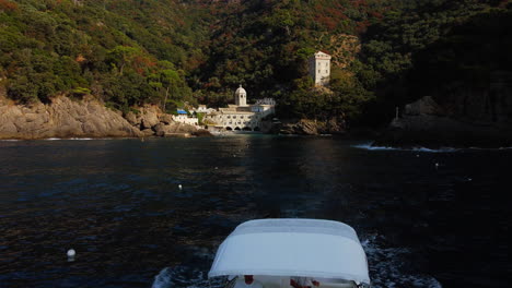 Drone-flying-over-a-boat-leaving-the-San-Fruttuoso-abbey,-in-sunny-Italy