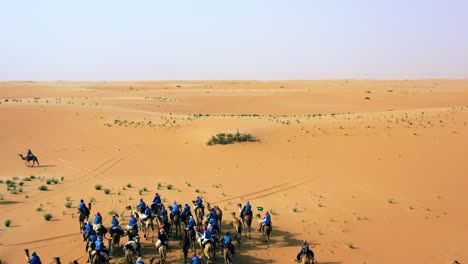 Aerial-view-around-a-camelcade-desert-safari,-Arabs-riding-on-a-endless-wasteland,-sunny-evening,-in-Saudi-Arabia---orbit,-drone-shot