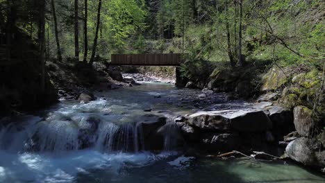 4K-Aerial-Drone-Flight-above-river-with-small-waterfall-towards-bridge-surrounded-by-forest