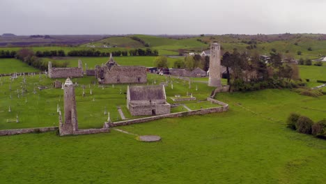 Dynamische-Luftaufnahme-Nähert-Sich-Alten-Runden-Türmen-Und-Gebäuden-In-Clonmacnoise