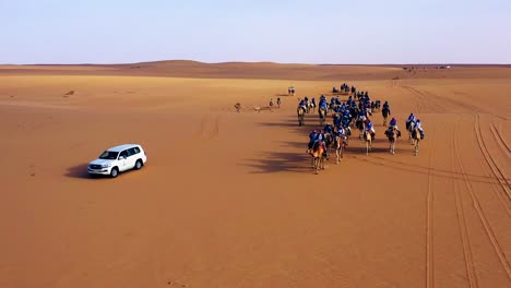 Aerial-view-around-a-camel-caravan,-Nomads-riding-camels-on-the-Arabian-Desert,-golden-hour,-in-Saudi-Arabia---orbit,-drone-shot