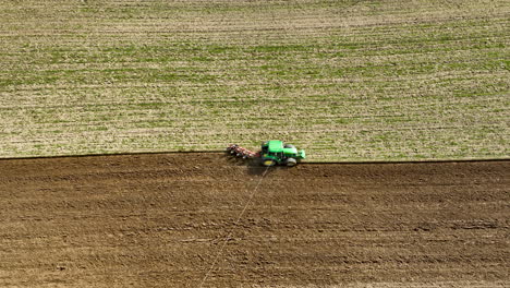 Primer-Plano-Aéreo-De-Un-Tractor-Verde-Con-Equipo-De-Arado-Trabajando-El-Suelo-En-Tierras-De-Cultivo,-Creando-Una-Línea-Clara-Entre-El-Campo-Arado-Y-El-área-De-Cultivo-Verde-Intacta