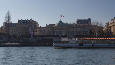 Bateau-Mouche-passing-by,-building-with-French-flag-in-background