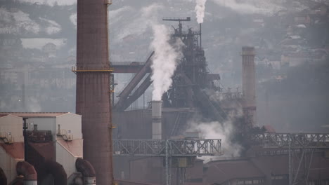 Industrial-smokestacks-emit-plumes-of-smoke,-with-a-backdrop-of-a-snowy-hillside-and-urban-landscape