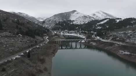 Frozen-mountain-range-peaks,-snowed-road-area-bridge-crossing-blue-river-aerial-drone-perspective-at-reservoir