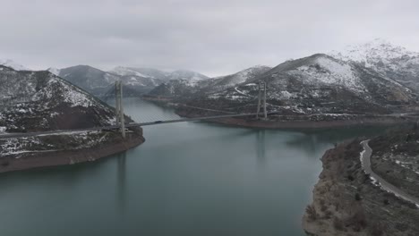 A-river-between-frozen-mountain-valley,-bridge-road-crossing-between-aerial-drone-infrastructure-reservoir-of-Barrios-de-Luna-in-Leon-,-Spain