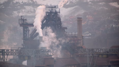 Rauch-Steigt-Aus-Industrieanlagen-Vor-Einer-Schneebedeckten-Bergkulisse-Auf-Und-Unterstreicht-Den-Starken-Kontrast-Zwischen-Industrie-Und-Natur
