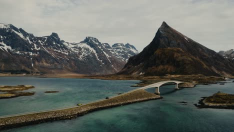 Driving-two-colorful-big-cars-over-a-bridge-in-beautiful-Lofoten-Islands-in-Northern-Norway-surrounded-by-mountains-peaks,-ocean,-beaches-and-snow-on-a-stormy-and-windy-day-with-some-sun