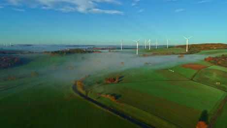 Aerial-view-over-rural-field-toward-sunlit-wind-turbines,-on-a-foggy-morning
