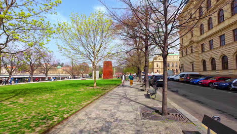 Slow-motion-shot-of-locals-walking-on-a-pavement-along-the-roadside-in-Prague,-Czech-Republic-on-a-bright-sunny-day