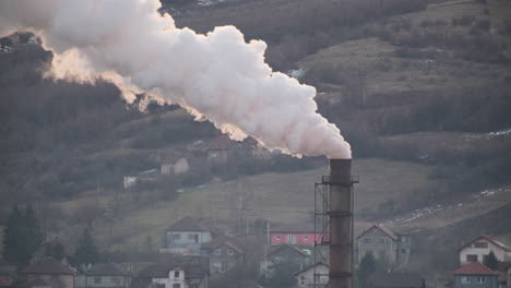 Thick-smoke-billows-from-a-large-industrial-chimney,-dominating-the-skyline-above-a-small-village-nestled-in-a-hilly-landscape