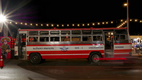 Timelapse-of-a-neighborhood-night-market-located-beside-the-road,-where-a-red-bus-is-parked-and-a-busy-traffic-passes-by-in-Chachoengsao-province-in-Thailand